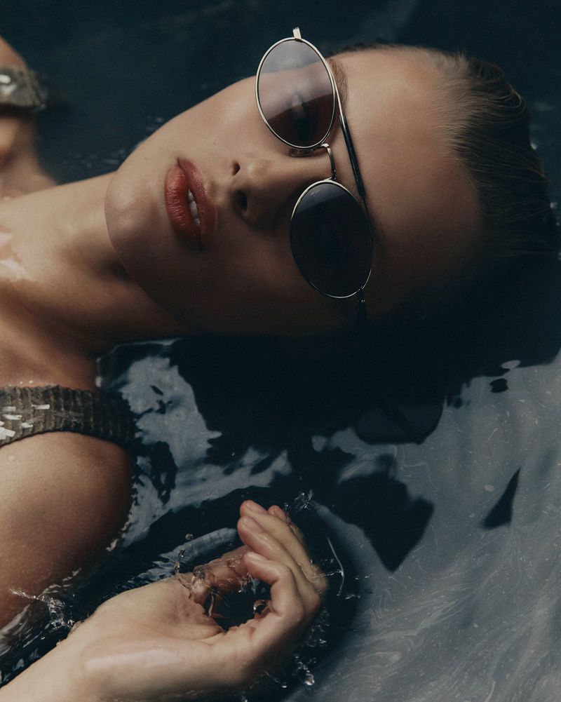 Close-up of a woman with sunglasses partially submerged in dark water, showcasing her face, hand, and reflective eyewear in soft lighting.