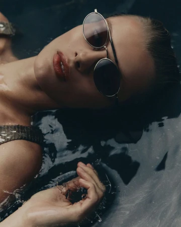 Close-up of a woman with sunglasses partially submerged in dark water, showcasing her face, hand, and reflective eyewear in soft lighting.