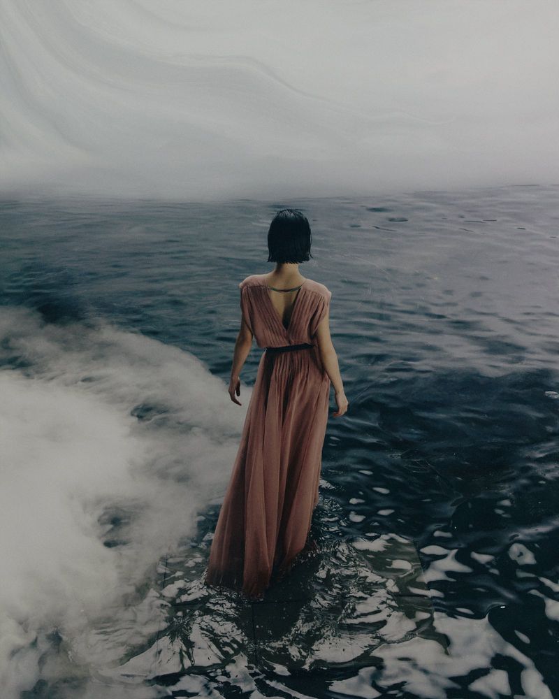 Model standing in shallow water wearing a long pleated pink dress seen from behind