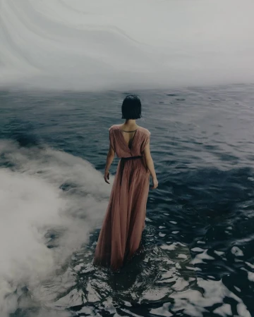 Model standing in shallow water wearing a long pleated pink dress seen from behind