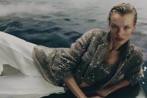 Kris Grikaite wearing a textured taupe jacket and white trousers reclining near water for Brunello Cucinelli