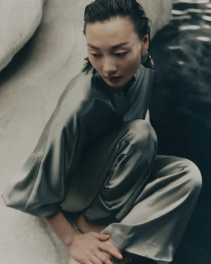 Model with slicked-back dark hair wearing olive green silk trousers and matching top crouching outdoors
