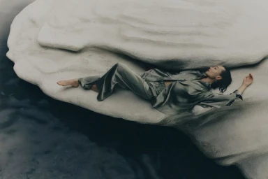 Model lying on a sculptural pale rock formation wearing a silky grey shirt and trousers