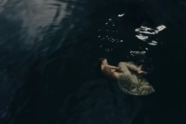 Model floating in dark water wearing a shimmering patterned skirt with soft highlights reflecting on the surface.