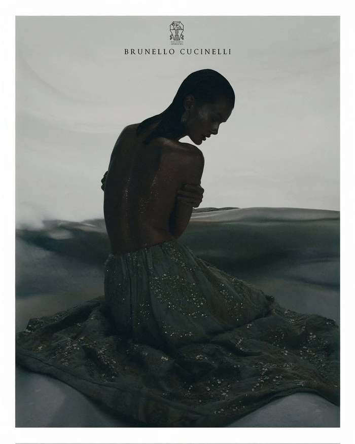 Model with slicked-back hair seated wearing a shimmering skirt against a muted grey background for Brunello Cucinelli