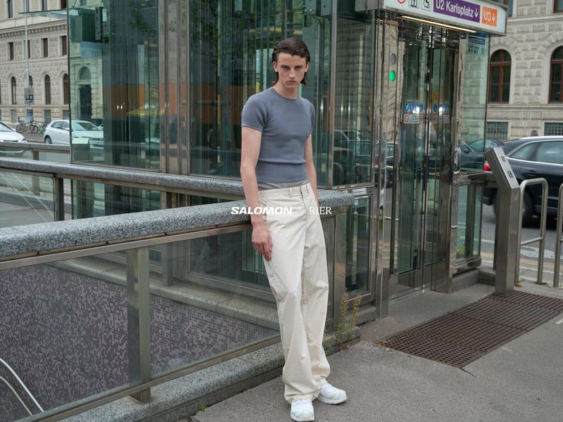 A slender man poses outdoors in a casual grey shirt and loose, light-colored trousers in an urban setting with a modern architectural design.