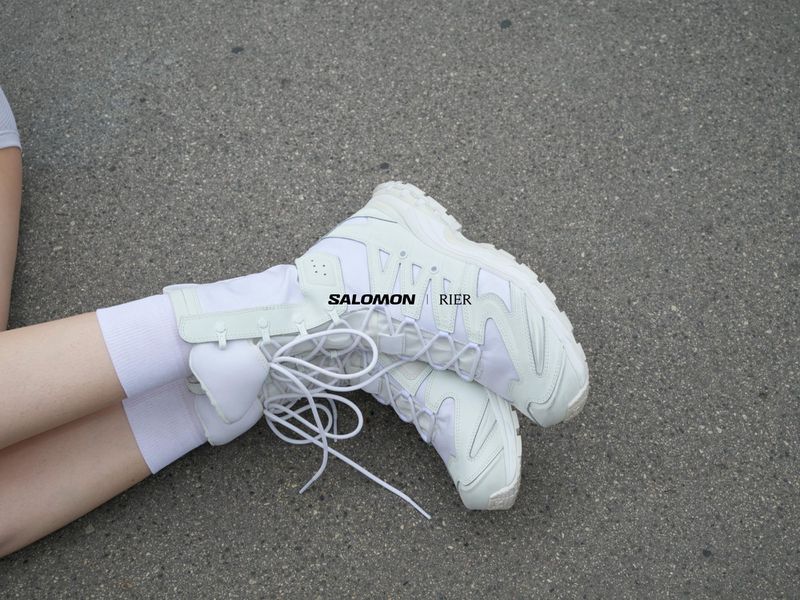 Close-up of white Salomon sneakers and socks, with a person's legs on a gray asphalt surface. Brand logos are clearly visible.