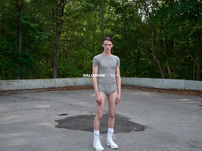 A slender adult male poses outdoors in matching gray shorts and tee with white sneakers, on pavement with trees in the background.