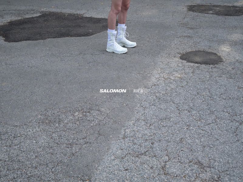 Person stands on cracked asphalt wearing white Salomon boots and socks, against background of pavement, puddles, and text logos.