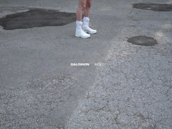 Person stands on cracked asphalt wearing white Salomon boots and socks, against background of pavement, puddles, and text logos.