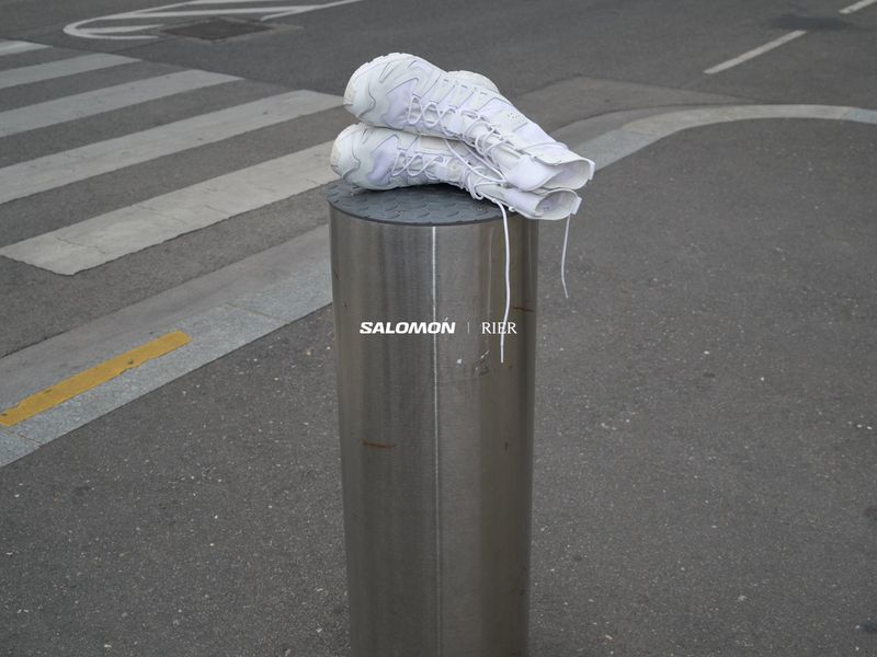 Two white Salomon sneakers placed atop a reflective metal bollard in a street setting, with a pedestrian crossing in the background.