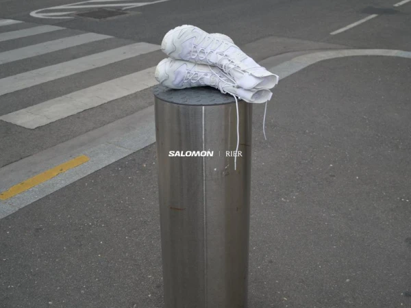 Two white Salomon sneakers placed atop a reflective metal bollard in a street setting, with a pedestrian crossing in the background.