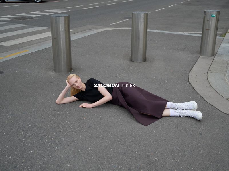 Woman in black top and brown skirt laying on city street with metal bollards and vehicle in background posing for fashion editorial.