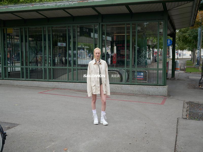 An androgynous model poses in a city street in a beige jacket, visible brands Salomon and Rier. Subway entrance and sidewalk.