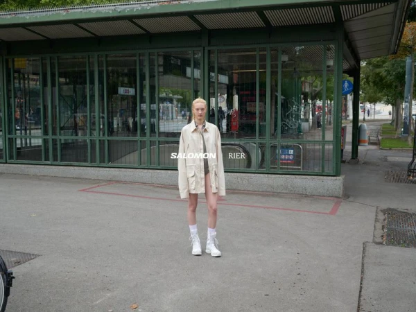 An androgynous model poses in a city street in a beige jacket, visible brands Salomon and Rier. Subway entrance and sidewalk.