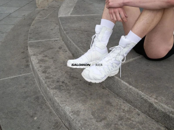 Person in white Salomon and Rier collaboration footwear sitting on concrete stairs, with a neutral expression, and minimal fashion style.