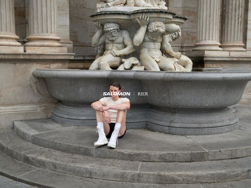 A person sits on stone steps near a fountain with sculptural figures, wearing white boots, set against an outdoor environment.