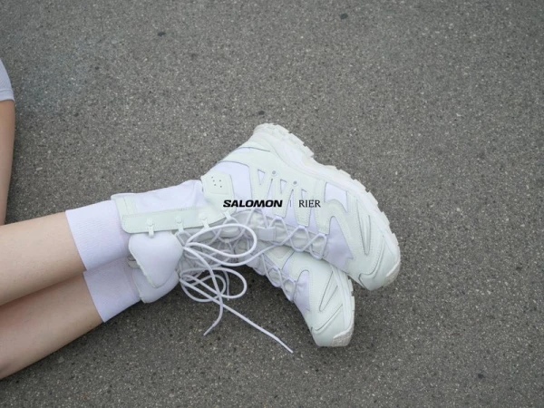 Close-up of white Salomon x Rier sneakers worn with white socks resting on a textured asphalt ground