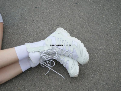 Close-up of white Salomon x Rier sneakers worn with white socks resting on a textured asphalt ground