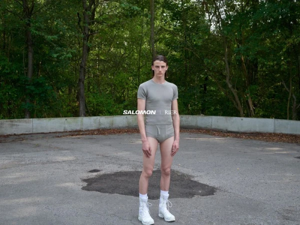 Model standing outdoors wearing a grey t-shirt, grey briefs, and white boots in a paved lot surrounded by trees.
