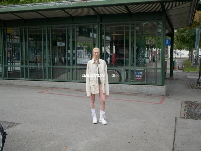 Model standing outdoors wearing an off-white multi-pocket jacket and white sneakers in front of a green framed pavilion.