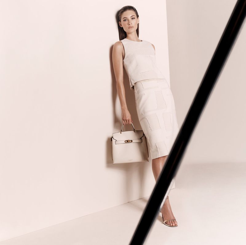 Elegant brunette woman in a cream top and skirt poses with a matching handbag in a bright photo studio with neutral tones.