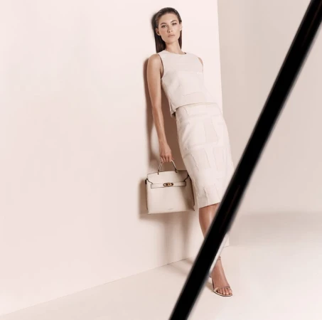 Elegant brunette woman in a cream top and skirt poses with a matching handbag in a bright photo studio with neutral tones.
