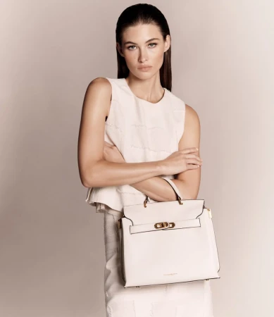 Elegant adult woman poses with an ivory satchel bag in a studio setting, showcasing a sleeveless top and knee-length skirt.