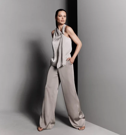 Full-length shot of a beautiful adult woman posing in a studio wearing a shiny halter top and wide-leg trousers, against a gray backdrop.