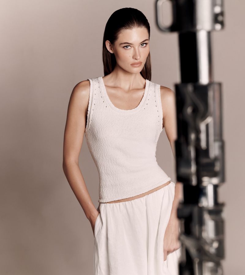 Woman poses in white tank top and pants against a neutral background, showcasing fashion apparel in a studio setting with soft lighting.