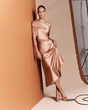 Elegant adult female poses in a shiny satin dress and high heels in a studio with an orange wall and film equipment in the background.