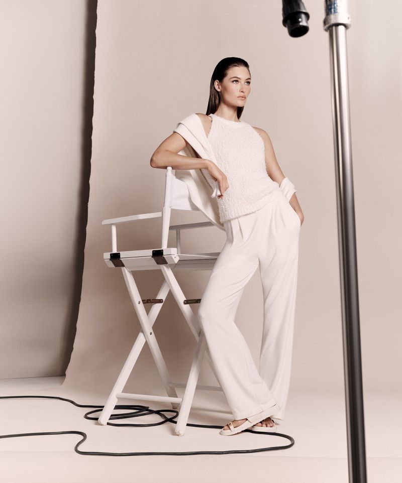 Brunette woman in all-white fashion poses in photo studio with director's chair and film lighting; stylish, elegant, and modern.