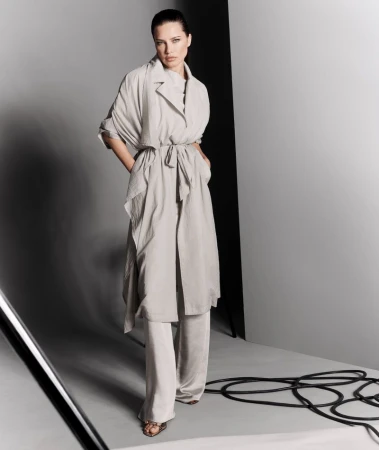 Woman poses in a light grey belted trench coat and matching trousers in a studio with black cables and white light reflector.