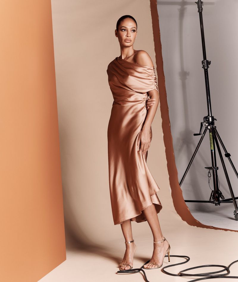 Fashion photography of a woman in a flowing gold dress, standing in a studio with a minimalist set and soft lighting. 