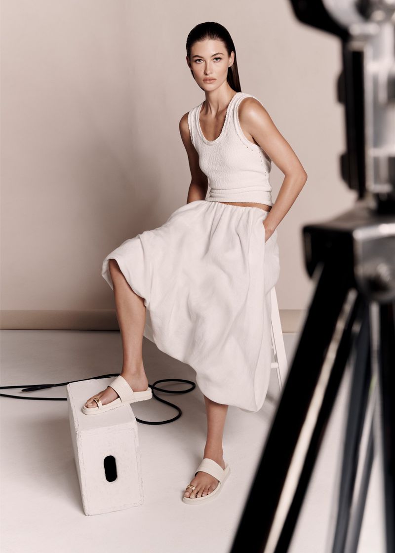 A fashionable adult woman poses in a photo studio wearing a matching tank top and skirt, complemented by white sandals and neutral makeup.