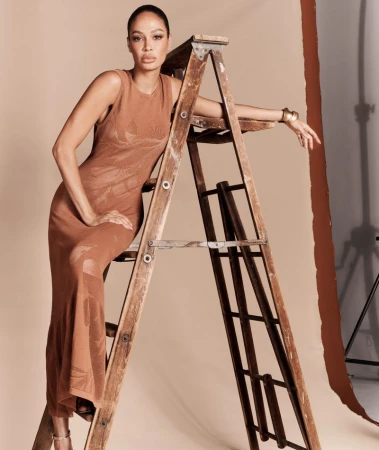 Joan Smalls posing on a wooden step ladder wearing a sleeveless brown knit dress for the Donna Karan campaign