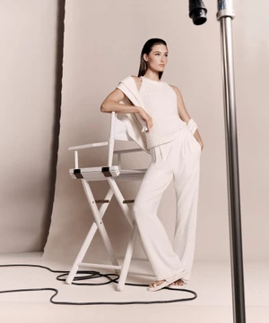 Model wearing white knit top and trousers leaning against a director chair in a studio setting