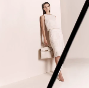 Grace Elizabeth leaning against a light wall in a cream top and skirt, holding a matching handbag