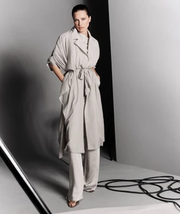 Model Adriana Lima in a belted grey coat and matching trousers standing against a studio backdrop.