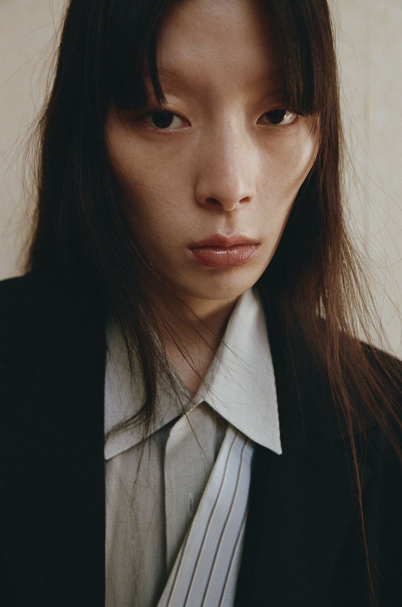 Close-up portrait of an East Asian woman wearing a white collared shirt and black blazer against a plain wall.