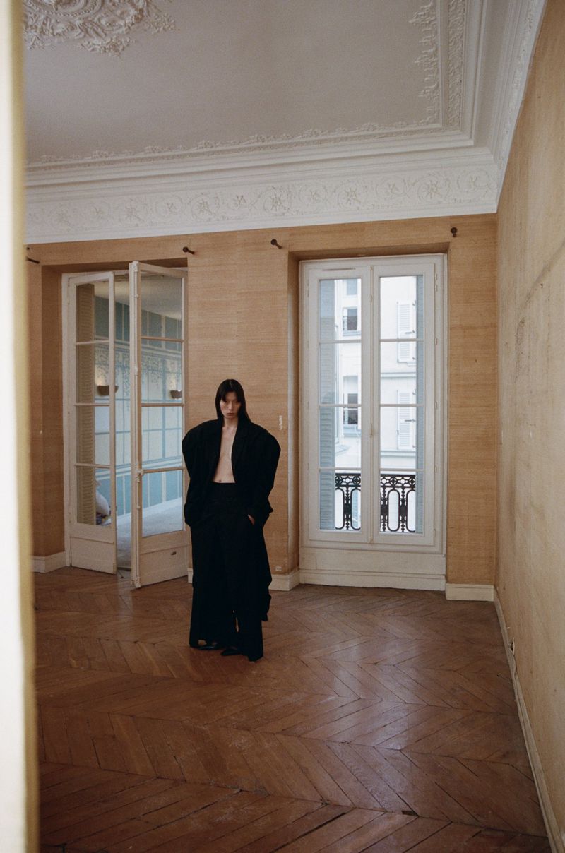 Indoor fashion shot of a slender woman in a black oversized suit, standing in an empty room with neutral walls and hardwood floors.
