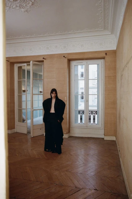 Indoor fashion shot of a slender woman in a black oversized suit, standing in an empty room with neutral walls and hardwood floors.