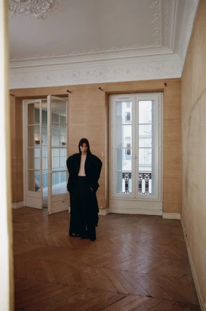 Indoor fashion shot of a slender woman in a black oversized suit, standing in an empty room with neutral walls and hardwood floors.