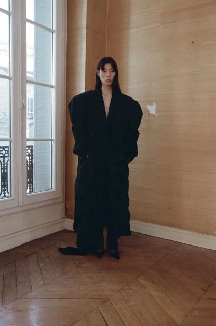 A woman with straight dark hair poses in an oversized black coat in a room with parquet floors and natural window light.
