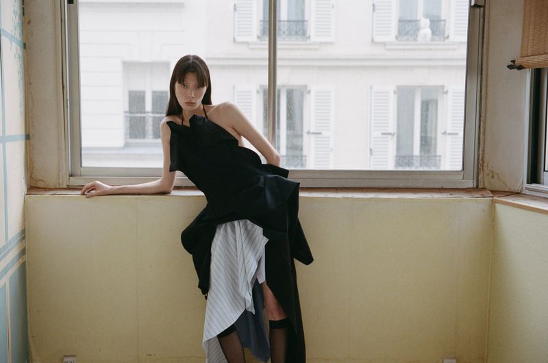 Asian woman in a black high-fashion dress with striped fabric sits by a bright window with a building view in an indoor setting.