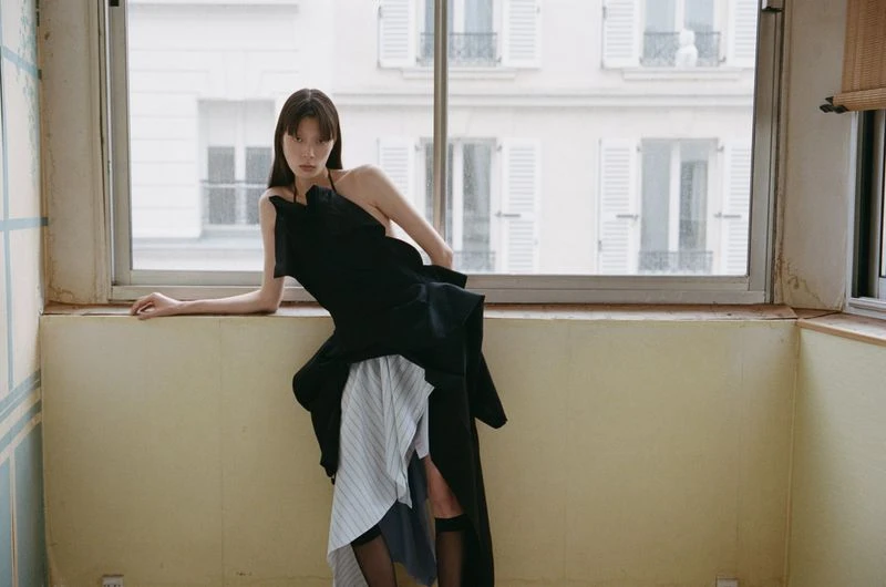 Asian woman in a black high-fashion dress with striped fabric sits by a bright window with a building view in an indoor setting.