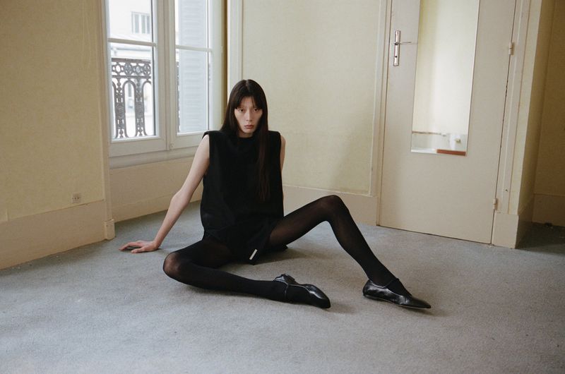Fashion photography of a slender woman in black clothing and tights, posing indoors on a carpet, with windows and a door visible.