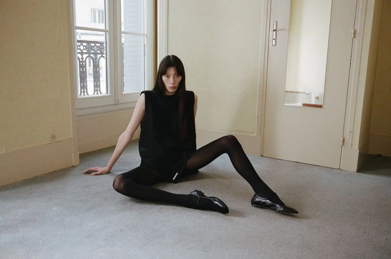 Fashion photography of a slender woman in black clothing and tights, posing indoors on a carpet, with windows and a door visible.