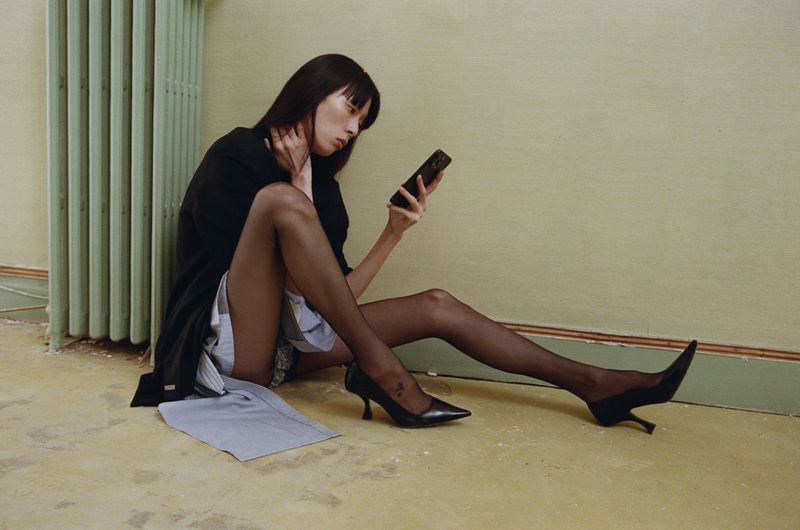 An Asian woman in a black jacket sits on the floor, holding a phone. She's wearing sheer tights and black high heels in an indoor setting.