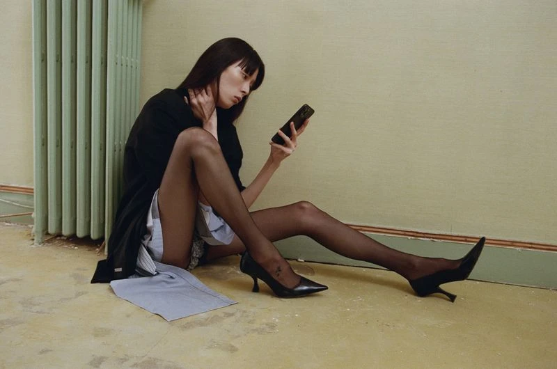 An Asian woman in a black jacket sits on the floor, holding a phone. She's wearing sheer tights and black high heels in an indoor setting.